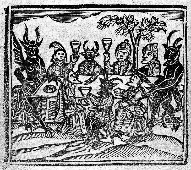 an old-style woodcut image of medieval people sat around a table with devils raising glasses in toast